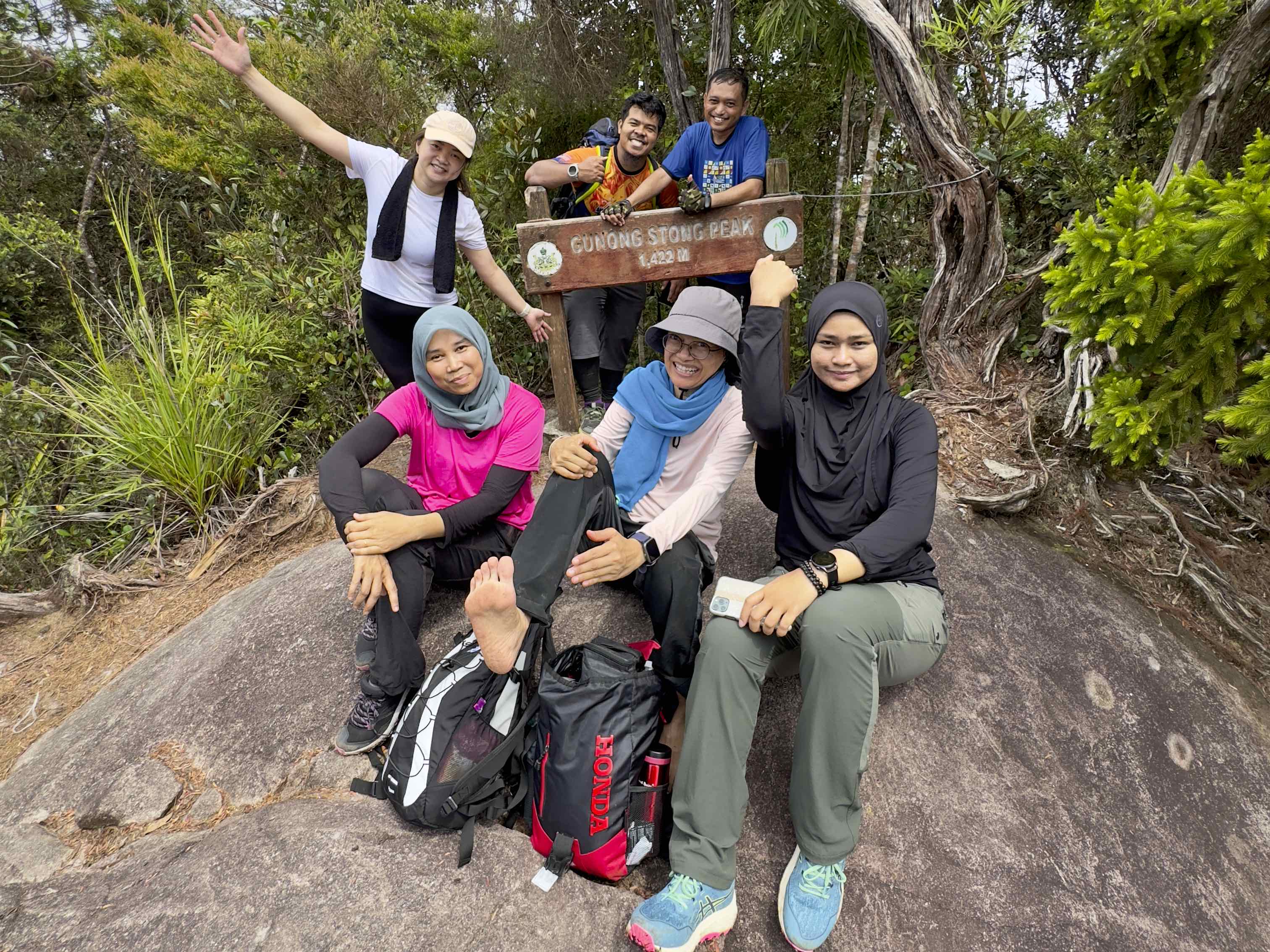 A group of five hikers celebrating at Gunun Stong Peak, with a wooden sign indicating the peak's height. They are posing on a rocky surface, surrounded by lush greenery.