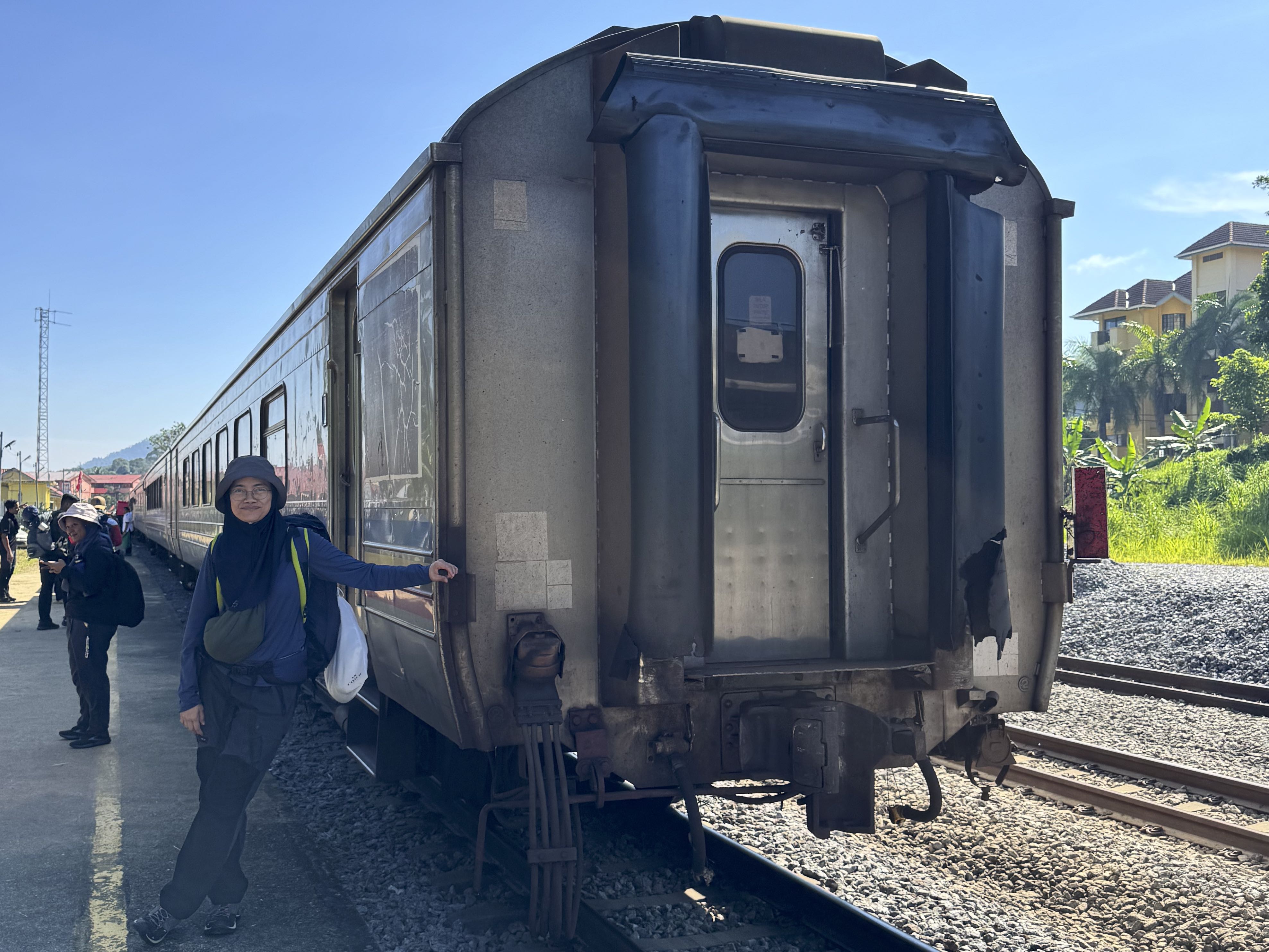 A person poses beside a train car at a station, with a bright blue sky and green vegetation in the background.