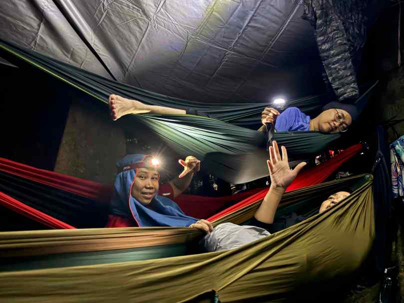 Three people relax in colorful hammocks indoors, wearing headlamps and smiling, with a tent-like structure above them.