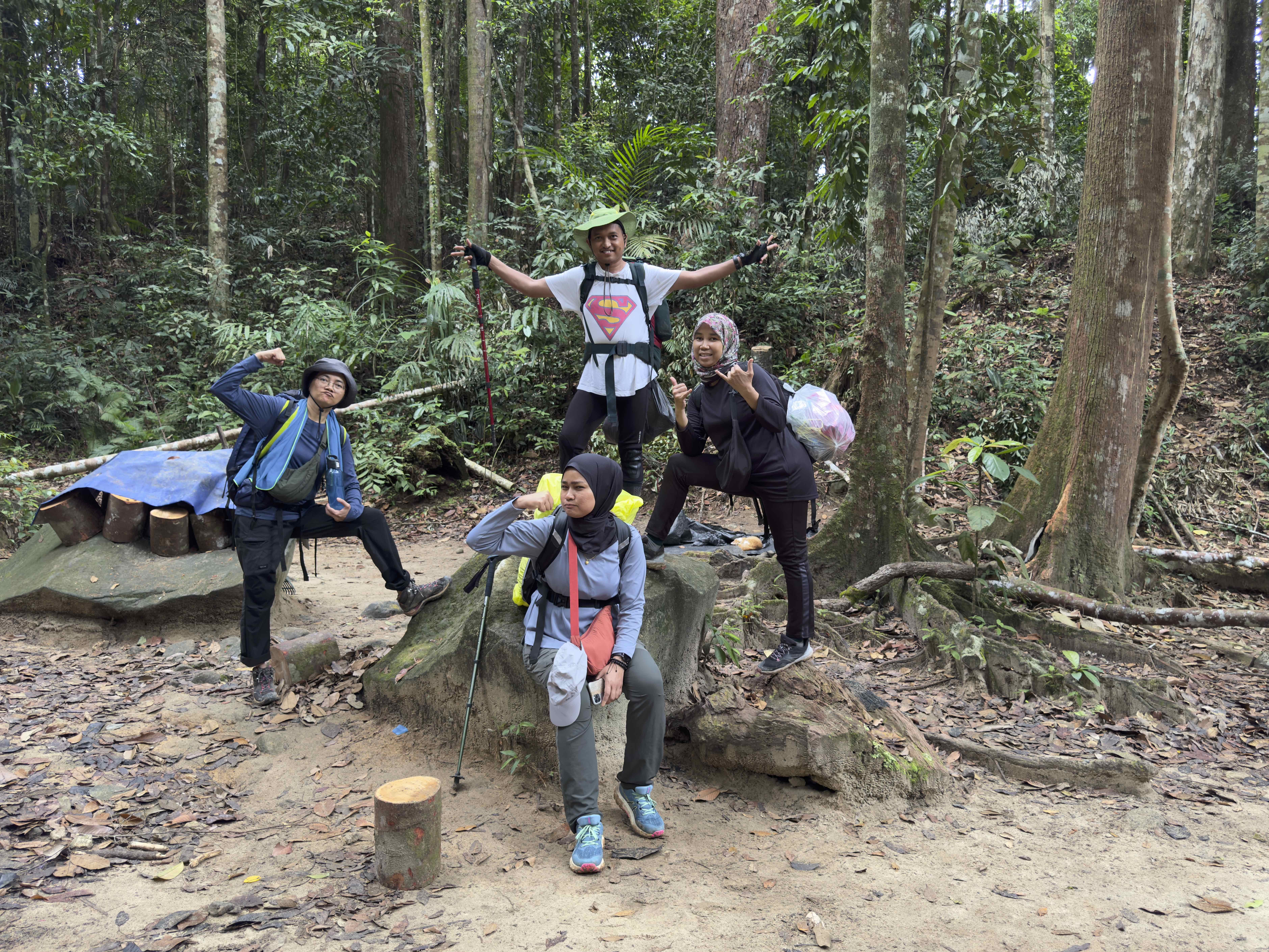 A group of four people posing enthusiastically in a forest setting, with one person flexing their muscles, another holding trekking poles, and the others making playful gestures. The area is surrounded by lush greenery and fallen leaves.