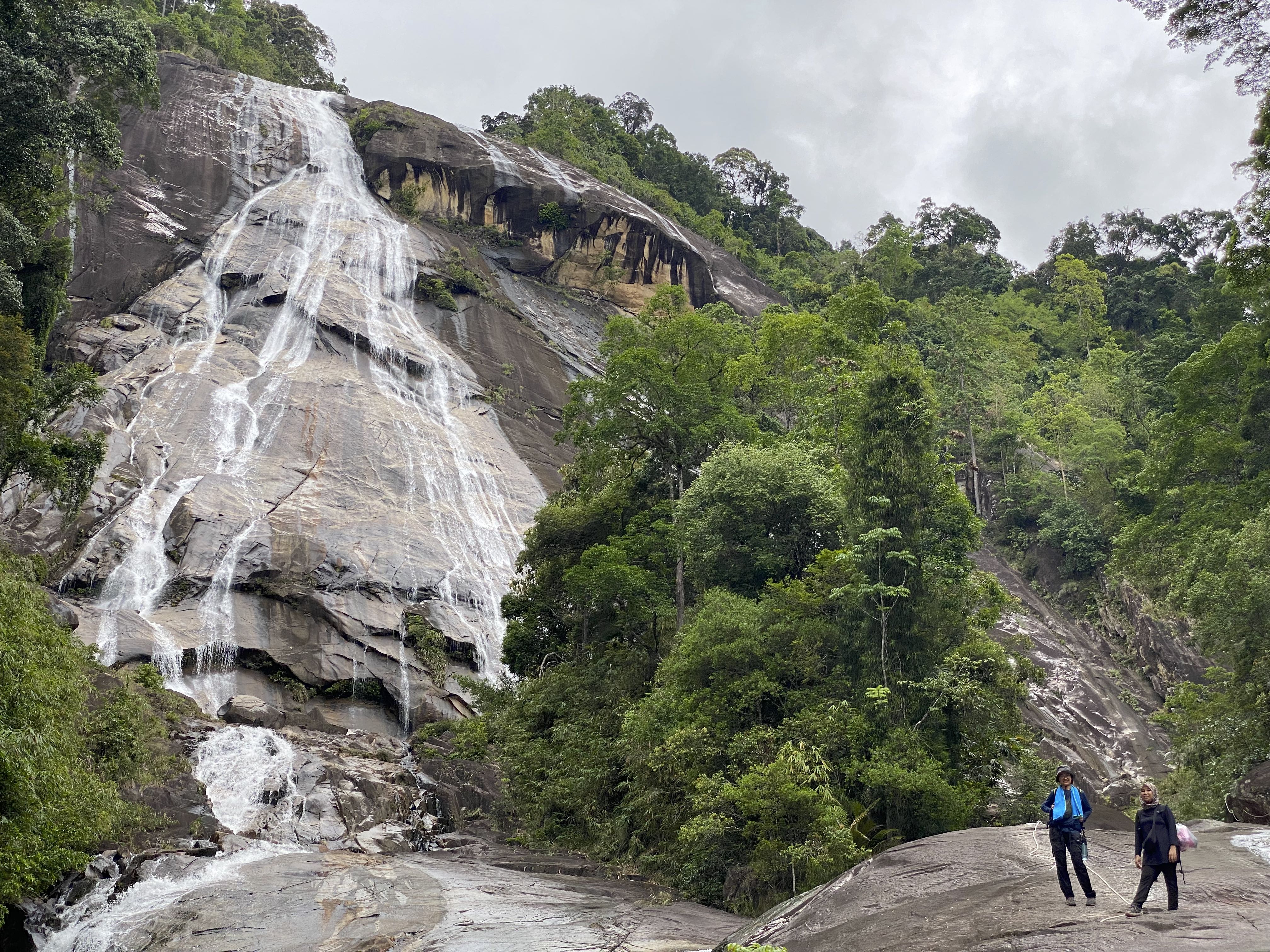 Two hikers standing on rocky terrain in front of a waterfall cascading down a steep cliff surrounded by lush greenery.