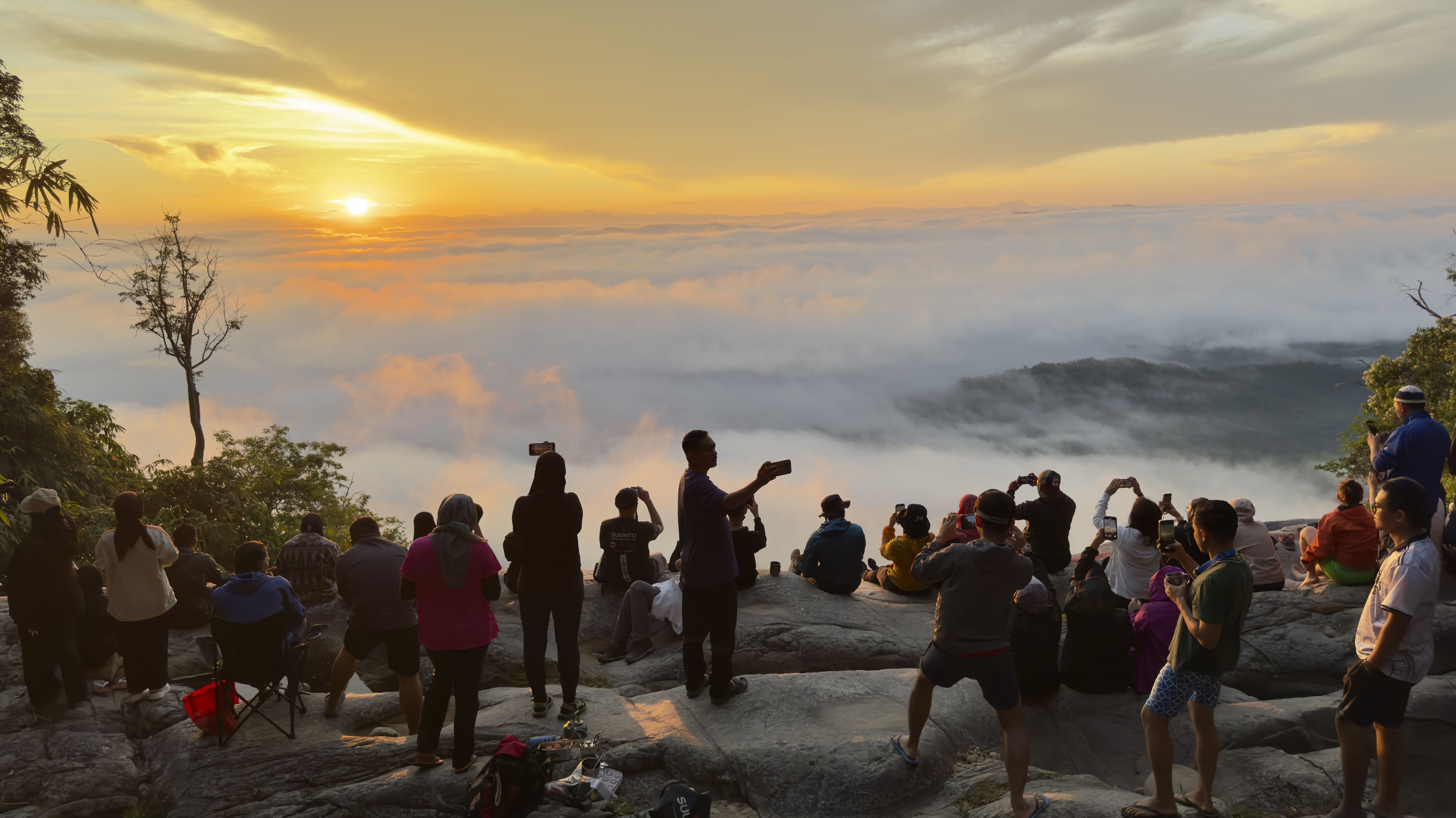 A crowd of people gathered on a rocky outcrop, watching a vibrant sunrise above a sea of clouds, with many individuals taking photos.