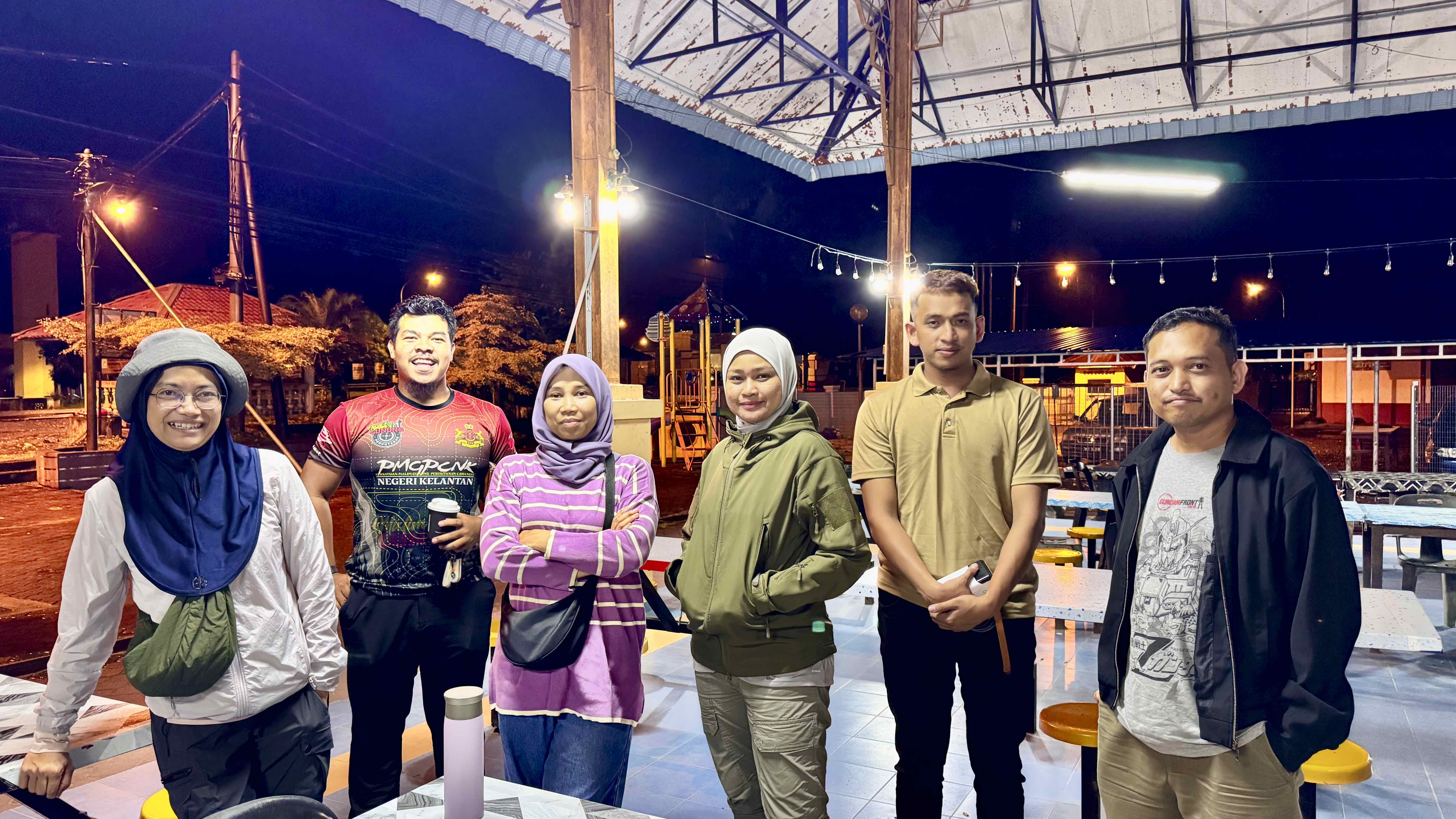 A group of six individuals standing together outdoors at night, with colorful lighting and a playground in the background.