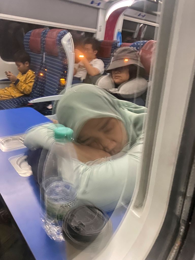 A woman wearing a light green headscarf sleeps on a train with her arms crossed. In the background, a child in yellow pajamas looks at a device, while another person with a hat rests with a neck pillow. Various beverage containers are on the table in front.