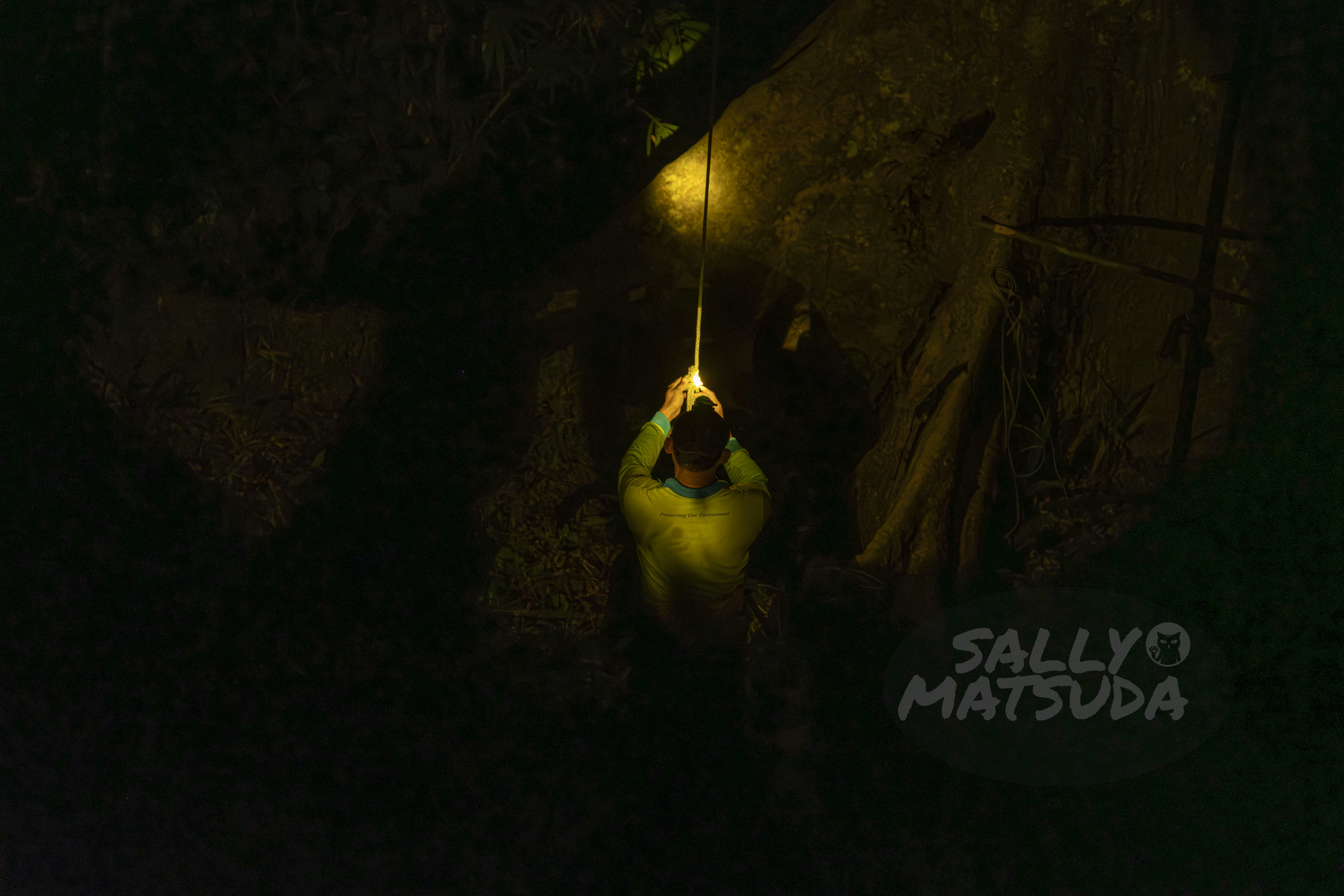 A person wearing a light-colored shirt is illuminated by a small light while working near a cliff or large rock in a dark forested area.
