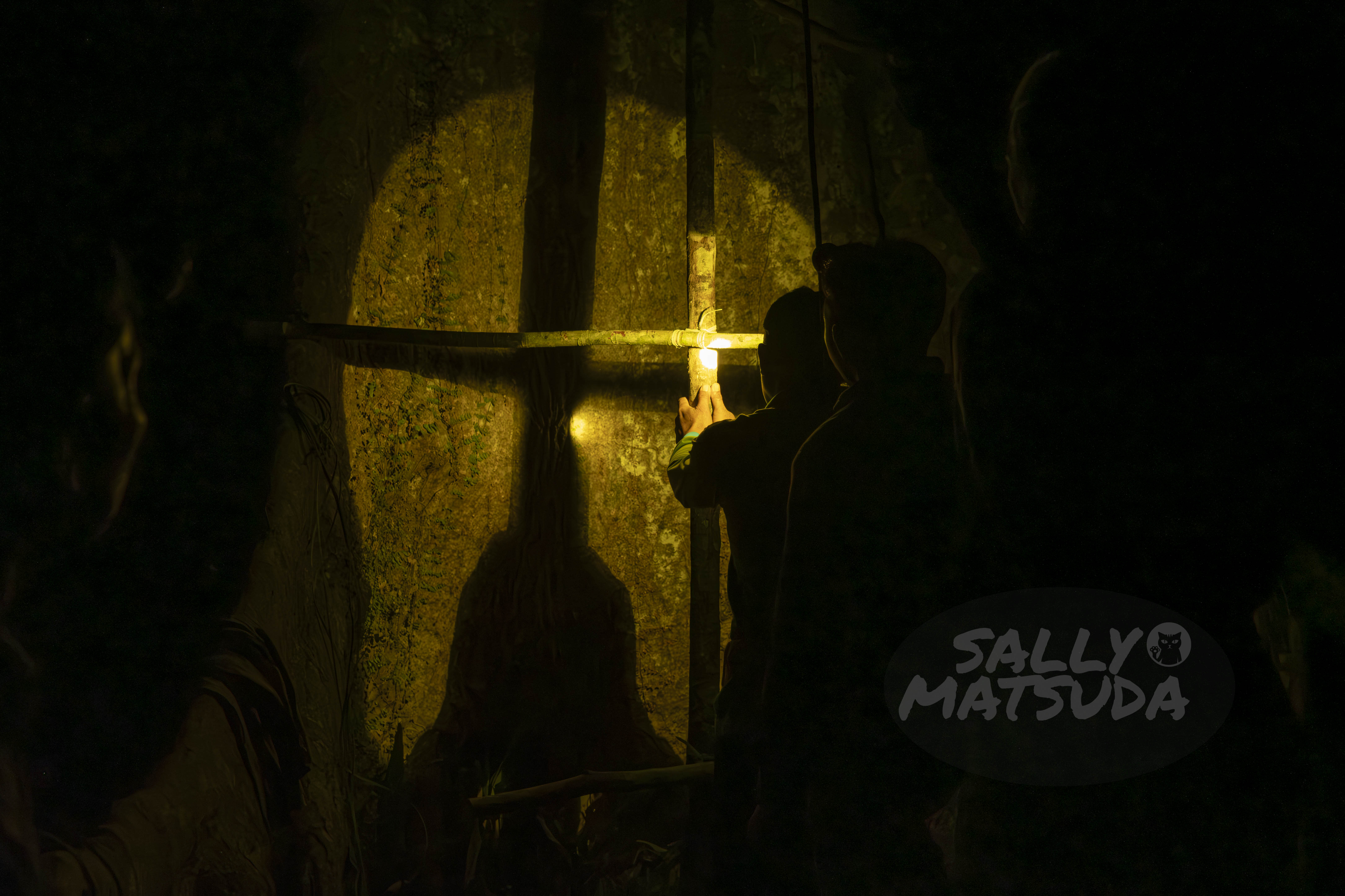 Figures in silhouette standing in a dimly lit area, holding a lantern that casts a warm glow on a textured surface behind them.