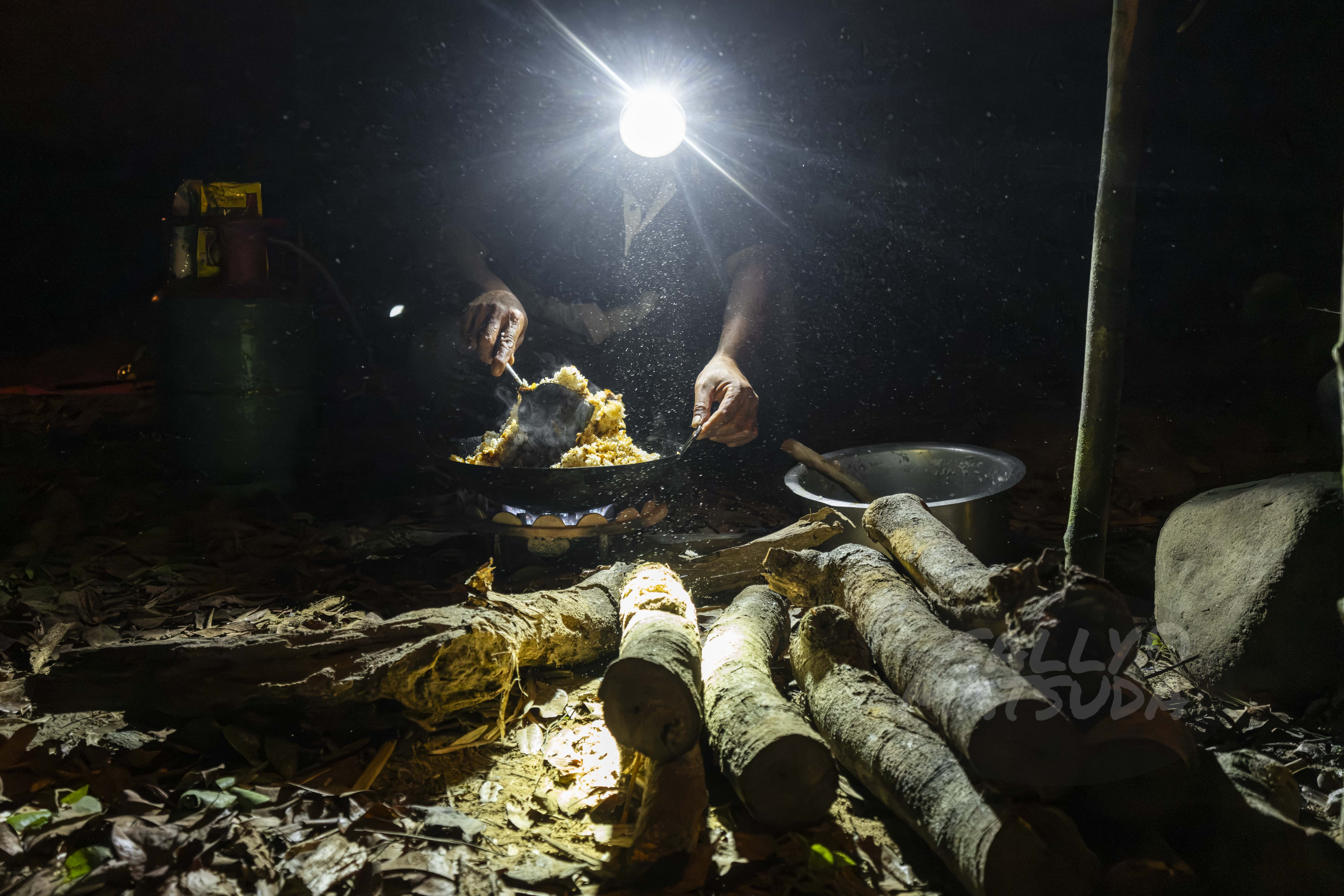 A person cooking outdoors at night, illuminated by a headlamp, with a frying pan filled with food, and logs scattered on the ground.