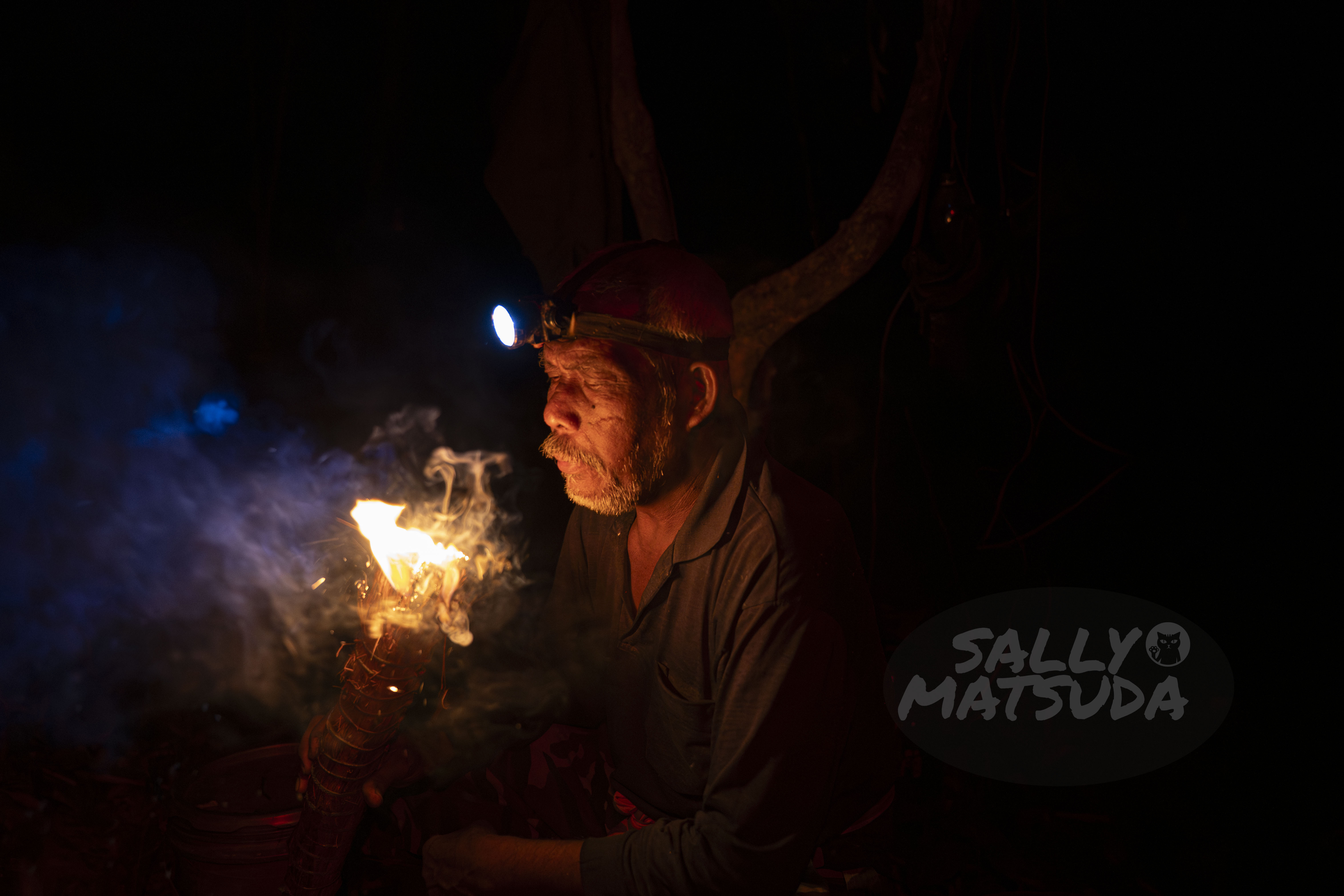 A man with a headlamp sitting in the dark, holding a flaming torch with smoke rising in the air.